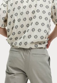 Camisa de manga corta en tejido texturizado color blanco roto con patrón de diamantes negros, combinada con pantalones gris claro con bolsillos traseros.