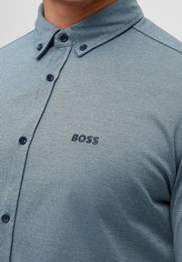 Vaaleansininen kuvioitu nappipaita, jossa on levitetty kaulus, tummansiniset napit ja brodeerattu "BOSS"-logo vasemmassa rinnassa.