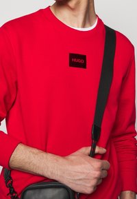 Homme portant un sweat-shirt rouge avec un patch logo "HUGO" noir, tenant une sangle de sac bandoulière noire sur sa poitrine.