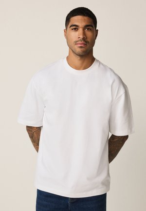 Muškarac s kratkom kovrčavom kosom i tetovažama na rukama, odjeven u jednostavnu bijelu široku majicu i tamnoplave traperice, stoji nasuprot bež pozadine.