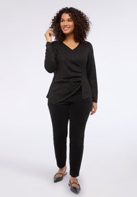 Mujer con cabello rizado que lleva una blusa negra de manga larga con destellos, pantalones negros y zapatos planos negros puntiagudos, de pie frente a un fondo blanco.