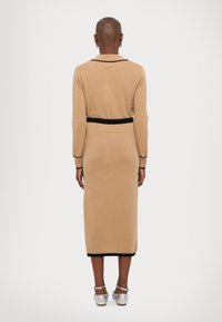 Beige knit dress with long sleeves, black trim at collar and waist. Slim fit with mid-calf length. Silver ankle-strap shoes are visible.