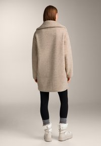 Beige oversized jas met een brede kraag, gestructureerde stof en doorgestikte schouders, gecombineerd met zwarte leggings en witte winterlaarzen.