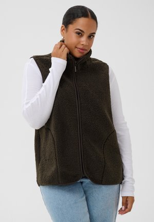 Jeune femme portant un gilet polaire marron foncé sur un haut blanc à manches longues et un jean bleu clair, tenant le col du gilet d’une main.