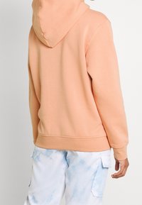 Broskvová mikina s kapucí na stahovací šňůrku a žebrovanými manžetami. Nošená s světle modrými batikovanými cargovými kalhotami s bočními kapsami.