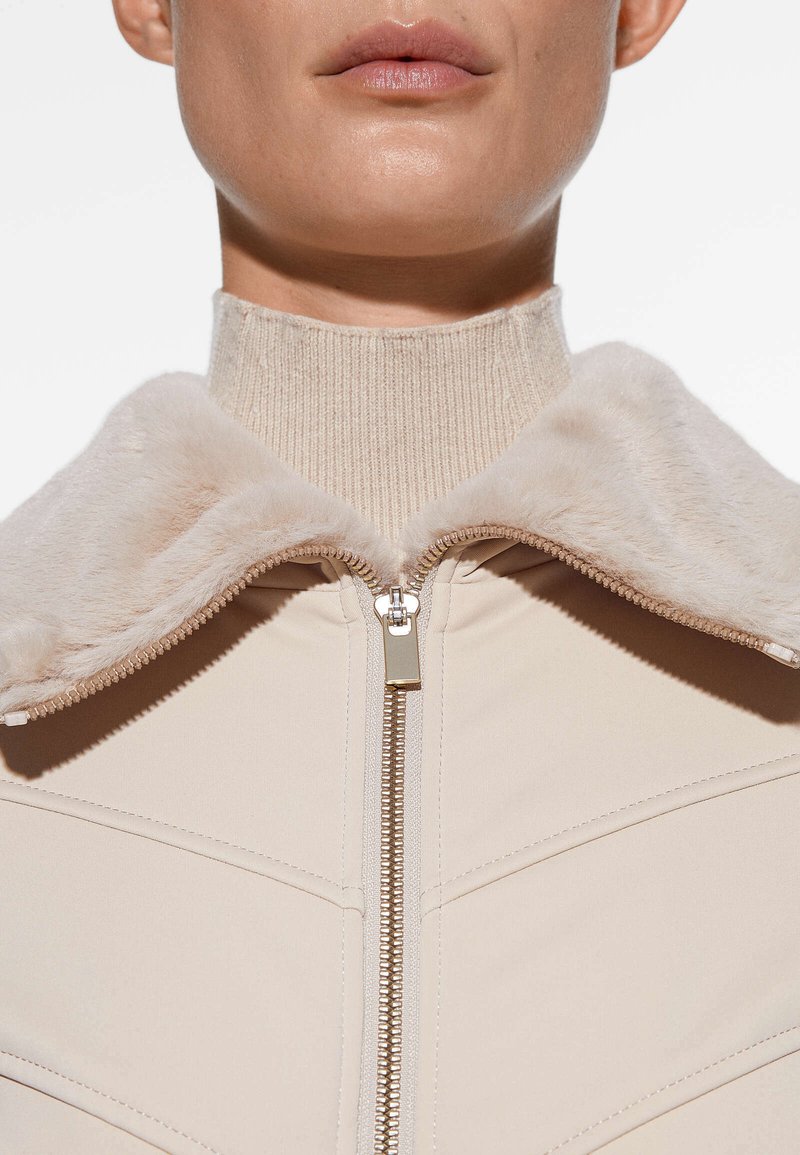 Chaqueta beige con un suave collar de tipo piel, cuello alto y cremallera plateada. Los paneles texturizados añaden interés visual al diseño aerodinámico.