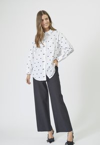 Camicia bianca con pattern di cuori neri, maniche lunghe e taschino sul petto, abbinata a pantaloni larghi scuri e scarpe con tacco nere.