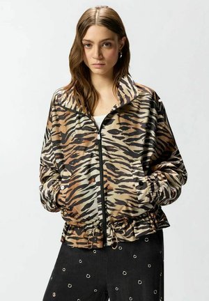 Giovane donna con lunghi capelli castani, indossa una giacca con stampa tigre con cerniera e pantaloni neri con occhielli argentati, mani nelle tasche, sfondo neutro.
