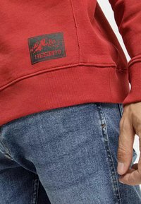 Czerwona bluza z ribbowanymi mankietami i dołem, z czarnym kwadratowym logo przedstawiającym górski wzór oraz napisem "1990". Noszona z niebieskimi dżinsami.