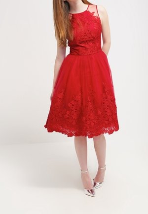 Mujer con vestido midi de encaje rojo sin mangas con bordado floral y tacones plateados con correa en el tobillo, de pie sobre un fondo liso.