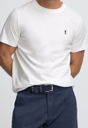 Mężczyzna w dopasowanej białej koszulce z małym, ciemnym logo polo z zawodnikiem na piersi, granatowych spodniach i plecionym granatowym pasku.