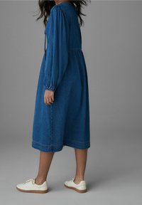 Robe midi en denim bleu profond, dotée de manches longues froncées et d'une taille cintrée. Associée à des baskets blanches et un ourlet texturé.