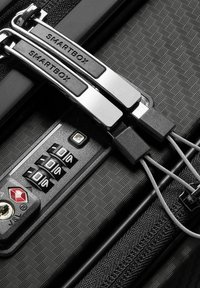 Gros plan d'une valise noire texturée avec serrure à combinaison TSA et deux tirettes de fermeture éclair argentées étiquetées "SMARTBOX" sécurisées par un cadenas à câble.