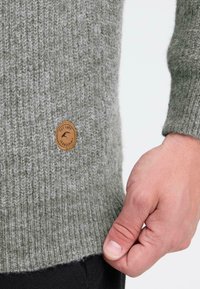 Hallitus halli kudum, ribatud tekstuuriga ja pruuni nahketegevusega sildiga külje peal. Käsi puudutab kergelt äärt. Reguleeritav randmeava.