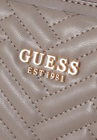 Materiale sintetico grigio trapuntato con logo dorato "GUESS EST 1981". Presenta cuciture in rilievo e una texture liscia.