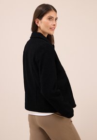 Donna con capelli castani che indossa un giubbotto nero strutturato sopra una maglietta bianca e pantaloni beige, in piedi davanti a uno sfondo beige uniforme.