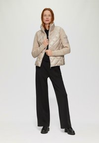 Beige gewatteerde puffer vest met een hoge kraag, geribbelde beige mouwen en een ritssluiting. Draag het over een zwarte top en combineer het met zwarte broeken.