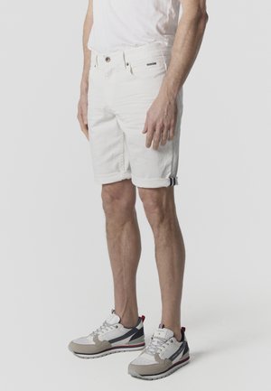 Mann trägt hochgekrempelte weiße Jeansshorts, weißes T-Shirt und weiß-graue Turnschuhe und steht vor einem einfarbigen Hintergrund.