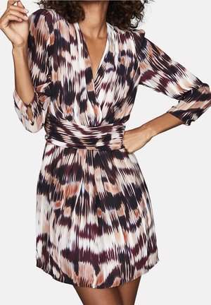 Kobieta w długiej sukience na pasku z długim rękawem, głębokim dekoltem w kształcie litery V oraz abstrakcyjnym wzorem w odcieniach brązu, czerni i bieli, pozująca z ręką na biodrze.