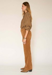 Blouse à carreaux marron avec des manches bouffantes, associée à un pantalon en suède beige et des bottes à bout pointu marron. Les accessoires incluent des bijoux en argent.