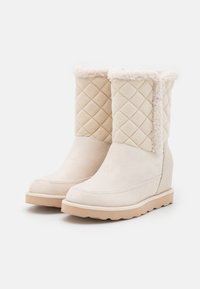 Stivali invernali beige con tomaia trapuntata, morbida fodera in shearling, tacco a zeppa e suola in gomma su sfondo bianco.