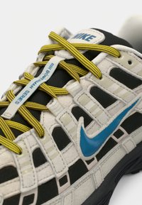 Tuva skats uz Nike sporta apavu ar bēšiem un melniem paneļiem, zilu zīmuli, dzeltenām svītrām auklām un tekstu "BMGERMAN SERIES" uz auklu turētāja.