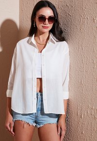 White linen shirt with long sleeves and button front, paired with a white crop top and denim shorts. Sunglasses and gold jewelry are worn.