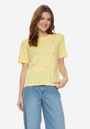 Młoda kobieta uśmiechająca się, ubrana w żółtą koszulkę w wielokolorowe kropki oraz jasnoniebieskie dżinsy z wysokim stanem, stojąca na białym tle.