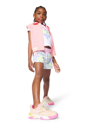 Young girl with braided hair wearing a pink sleeveless vest, white graphic tee, floral shorts, and beige sneakers with pink accents.