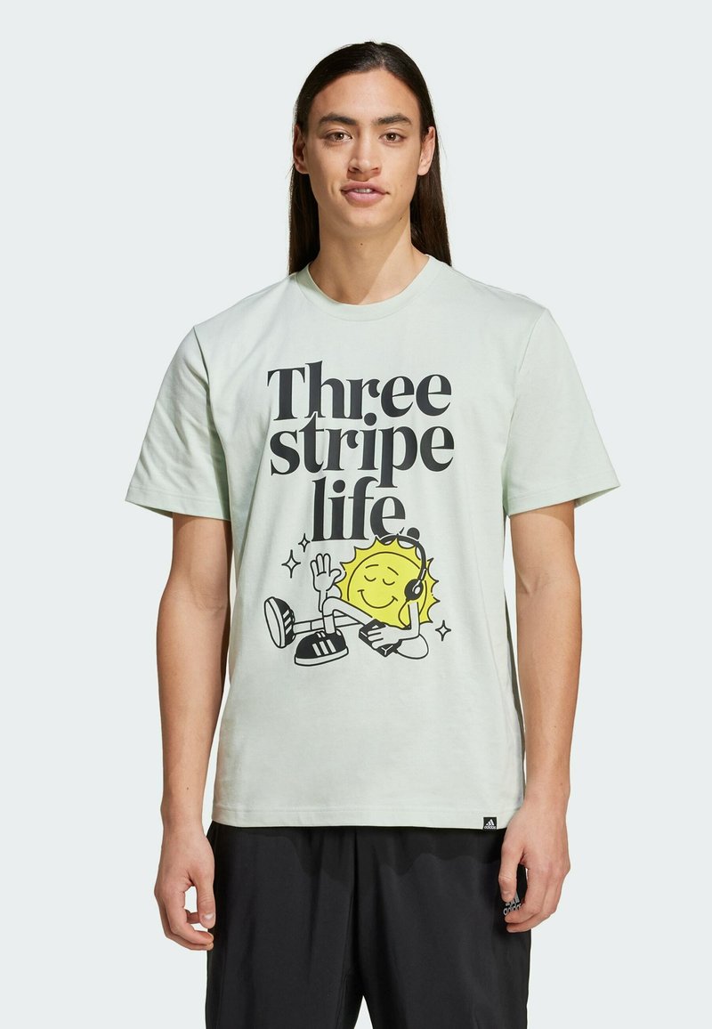 T-shirt di cotone verde chiaro con testo nero in grassetto "Tre strisce vita" e un personaggio sole dei cartoni animati con le cuffie al centro.