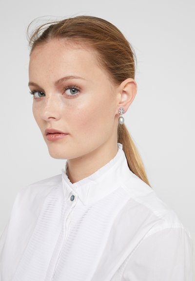 Cercei argintii cu accent de cristal și o perlă gri, purtați de un model cu păr deschis și cămașă albă cu guler.