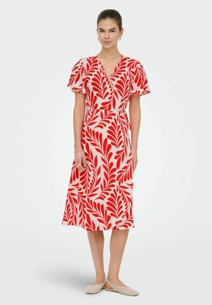 Žena stojaca, oblečená v krémových šatách po kolená s červeným lístkovým vzorom, s krátkymi volánovými rukávmi a výstrihom do V, doplnená béžovými pletenými balerínami.