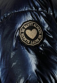 Giubbotto invernale navy con finitura lucida, dotato di una patch circolare nera ricamata con un design a forma di cuore e testo.