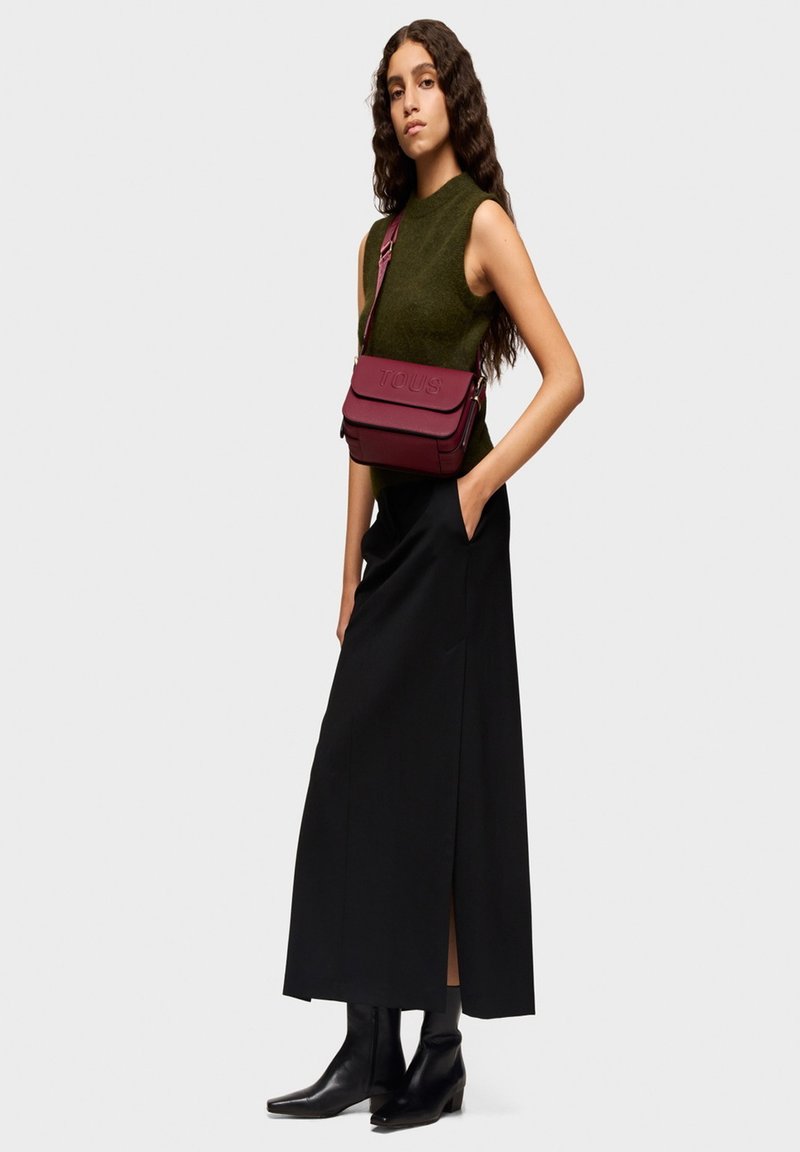 Falda maxi negra con una abertura alta, top sin mangas verde oliva y bolso cruzado de cuero burdeos con logotipo en relieve, combinado con botas negras de tobillo.