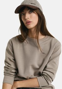 Lichtbruine sweatshirt met ribgebreide manchetten, voorzien van een klein geborduurd embleem. Grijze pet met een bruine klep en "gari" tekst aan de voorkant.