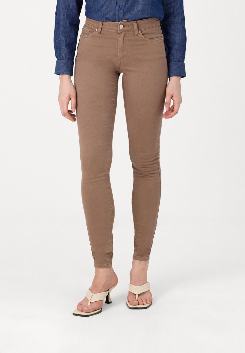 Personne portant un pantalon marron slim, une chemise bleue à manches longues et des sandales beiges à talons, debout devant un fond uni.