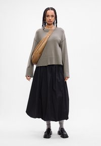 Oliwkowy sweter, czarna plisowana spódnica, szare skarpetki i czarne buty, z beżową torbą na ramię. Miękka tekstura i luźny krój.