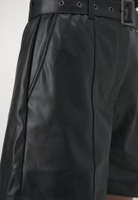 Pantaloni corti in pelle nera con una finitura testurizzata, dotati di una vita ampia, passanti per cintura e tasche laterali. Bottoni e cuciture sono visibili.
