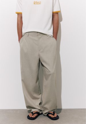 Mann trägt weite beige Hose, weißes T-Shirt mit dem Aufdruck „Kunst lebt auf den Straßen“ und schwarze Flip-Flops, steht mit den Händen in den Taschen.