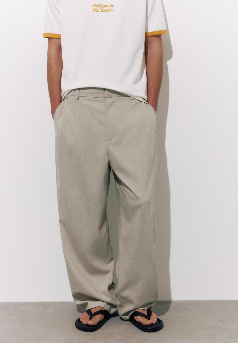 Homme portant un pantalon beige ample, un t-shirt blanc avec le texte « L'art vit dans les rues » et des tongs noires, debout les mains dans les poches.