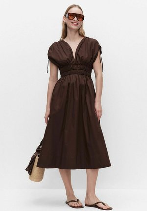 Vrouw draagt een donkerbruine midi-jurk met geplooide taille en mouwen, oversized zonnebril, teenslippers, en houdt een strohandtas met stoffen detail vast.