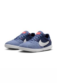 Kék velúr és hálós sportcipők fehér Nike logóval, piros részletekkel a nyelvén és perforált díszítéssel, texturált talppal rendelkeznek.