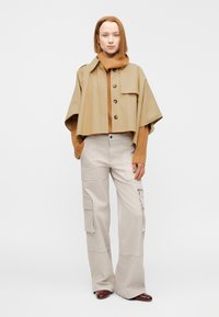 Veste courte beige clair avec boutons portée sur un pull marron côtelé, associée à un pantalon cargo gris clair avec poches latérales et ourlets.