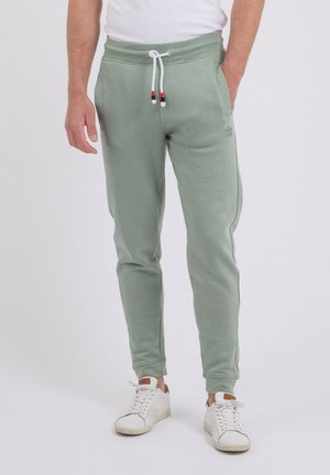 Homme portant un pantalon de jogging vert clair avec des cordons blancs et des baskets blanches, debout devant un fond uni.