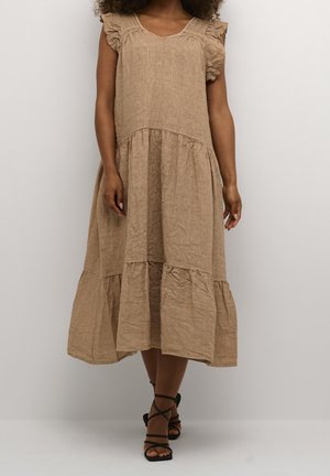 Kobieta w luźnej, beżowej sukience midi z warstwami, z falbanianymi, krótkimi rękawami typu cap i czarnych, rzemykowych sandałach, stojąca na jednolitym tle.
