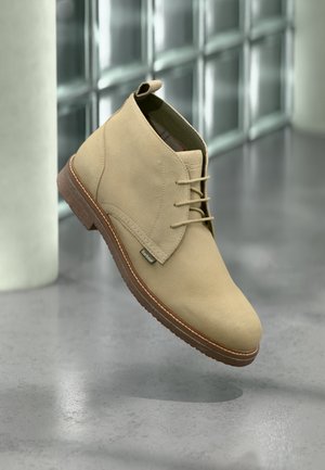Bottines en daim beige avec une texture lisse, un design à lacets et une semelle en caoutchouc marron avec un détail de couture contrastante.