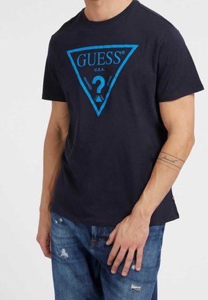 Homme portant un t-shirt GUESS bleu marine avec un logo triangle bleu vif et un jean bleu déchiré, tatouage visible sur l'avant-bras gauche.