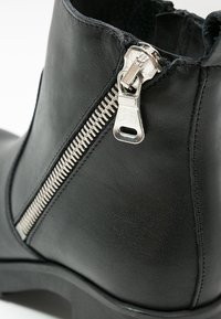 Botín de cuero negro con una cremallera de metal plateado, textura suave y punta redondeada. Presenta un detalle de costura a lo largo de la zona de la cremallera.