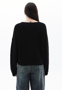 Maglione nero a maglia con vestibilità rilassata, scollatura ampia e orlo a coste, abbinato a jeans blu chiaro dal taglio morbido. Dettagli di texture sottili.