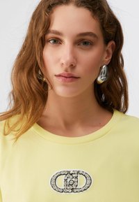 Jeune femme aux cheveux bruns ondulés portant des boucles d'oreilles argentées imposantes et un haut jaune clair avec un logo circulaire orné de bijoux sur la poitrine.